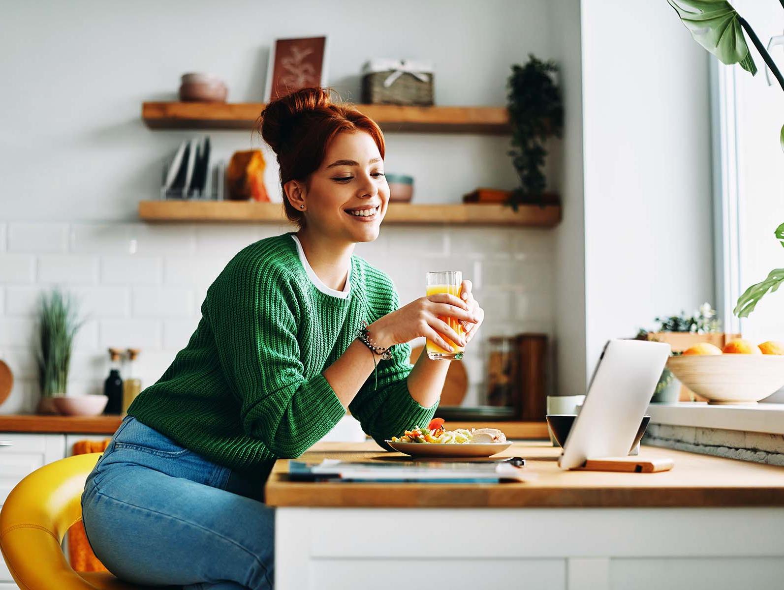 A smiling woman with a drink at a kitchen counter with a laptop.